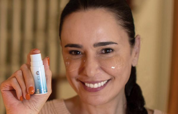 A brunette woman holding up an Eye Serum bottle while wearing Eye Patches under her eyes.